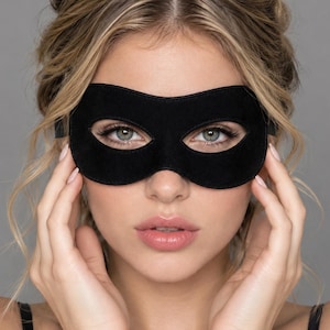 May include: A solid black eye mask featuring a smooth, matte finish and a contoured shape that covers the upper face. The mask has two oval-shaped cutouts for the eyes and is secured with a thin black strap extending from the sides.