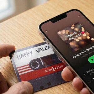 Peut inclure: Une carte rectangulaire avec un graphisme de cassette audio vintage portant le texte HAPPY VALENTIN et SCAN2PLAY, tenue à côté d'un smartphone affichant une application musicale avec une playlist intitulée Valentin's Romance et une icône cœur.