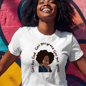 Può includere: Una t-shirt bianca a maniche corte con un'illustrazione grafica di una persona dalla pelle scura e una grande acconciatura afro. Il testo che circonda l'illustrazione recita, Girl got God. Girl got grace. Girl got goals.