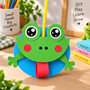 May include: A paper craft frog face with large white and black eyes, red circular cheeks, and a red tongue loop, mounted on a blue circular base. A yellow pencil extends vertically from the top of the green frog head, which features two small black nostrils.