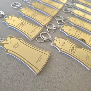 May include: A collection of gold-coloured acrylic keychains shaped like sports jerseys. Each keychain features a silver metal ring, engraved names, position abbreviations such as GD or WA, and the text Aggies Bulls 2026. The edges are white.