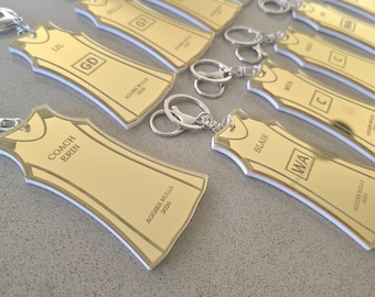 May include: A collection of gold-coloured acrylic keychains shaped like sports jerseys. Each keychain features a silver metal ring, engraved names, position abbreviations such as GD or WA, and the text Aggies Bulls 2026. The edges are white.