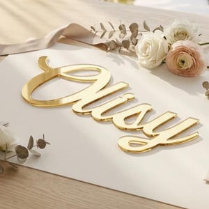 May include: A gold-coloured, reflective acrylic sign featuring the name "Nisy" in a cursive script font. The sign is placed on a white rectangular card, surrounded by light-coloured roses, eucalyptus sprigs, and several closed envelopes.