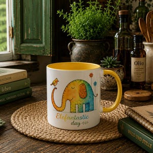 Könnte beinhalten: Ein weißer Keramikbecher mit gelbem Innenraum und Henkel, verziert mit einer bunten Aquarellillustration eines Elefanten mit einem kleinen Vogel auf dem Rüssel und dem Text Elefantastic day in mehrfarbiger Schreibschrift auf der Vorderseite.