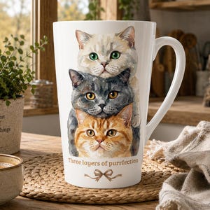 May include: A white ceramic latte mug featuring a watercolour illustration of three stacked cats in cream, grey, and orange colours. The text "Three layers of purrfection" is printed below the cats, accompanied by a small brown bow graphic.