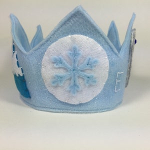 May include: A blue felt crown with a white snowflake and a blue and white castle design. The crown is decorated with glitter.