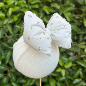 May include: A white headband with a large white bow decorated with silver beads. The headband is made of a soft, stretchy material and is perfect for adding a touch of elegance to any outfit.
