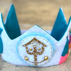May include: A white felt crown with a teal blue felt trim and a gold glitter design in the center. The crown has a circular design with a gold glitter symbol in the center.