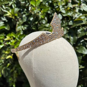 May include: A gold-colored tiara with a crown-like design. The tiara is encrusted with numerous small, sparkling crystals. The headpiece is displayed on a mannequin head against a backdrop of green foliage. This tiara is suitable for special occasions.