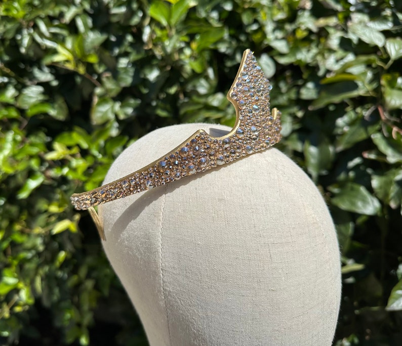 May include: A gold-colored tiara with a crown-like design. The tiara is encrusted with numerous small, sparkling crystals. The headpiece is displayed on a mannequin head against a backdrop of green foliage. This tiara is suitable for special occasions.
