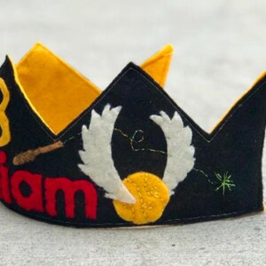 May include: A black felt crown with a yellow felt lining. The crown has a red felt inscription that says "William" and a yellow felt number "8". The crown also has a white felt design of a winged lightning bolt and a yellow felt circle.