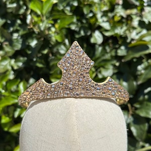 May include: A gold-toned tiara with a central diamond-shaped peak and a curved band. The tiara is encrusted with numerous clear, sparkling rhinestones. The tiara is displayed on a mannequin head against a backdrop of green foliage.