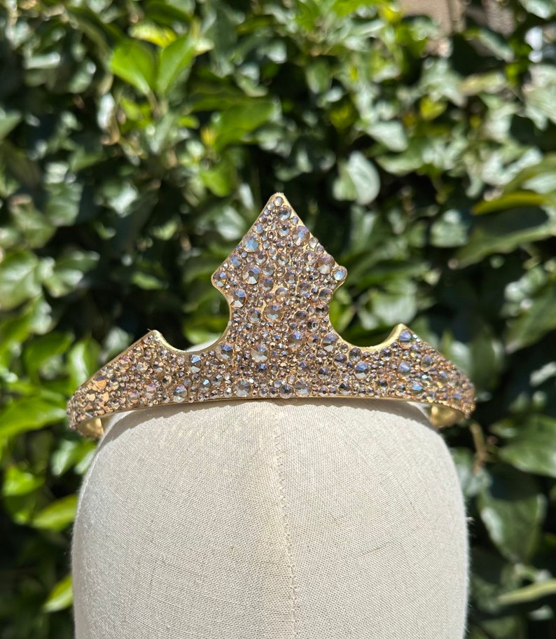 May include: A gold-toned tiara with a central diamond-shaped peak and a curved band. The tiara is encrusted with numerous clear, sparkling rhinestones. The tiara is displayed on a mannequin head against a backdrop of green foliage.