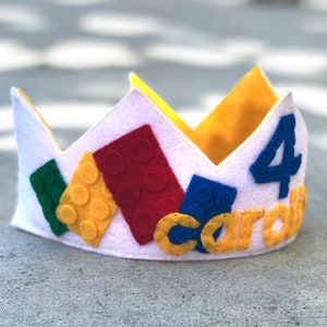 May include: A white felt crown decorated with colorful felt Lego blocks and the number 4. The crown also has the name "Carlos" written in yellow felt.