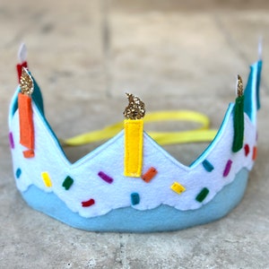 Può includere: Una corona di compleanno in feltro blu con candele gialle, arancioni e verdi e spolverata di coriandoli colorati. La corona ha un nastro giallo.