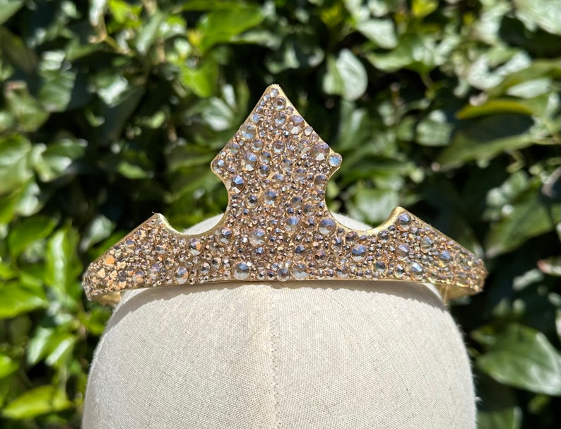May include: A gold-colored tiara with a central point and curved sides, encrusted with numerous clear, sparkling rhinestones. The tiara is displayed on a white mannequin head against a backdrop of green foliage.