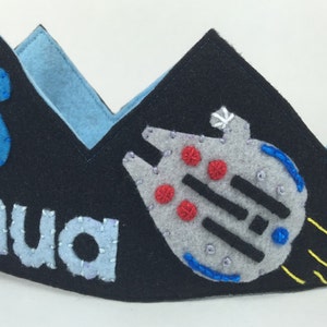 May include: A black felt crown with a blue felt number 6, the name "Joshua" in blue felt, and a gray felt spaceship with red and blue details. The spaceship has yellow felt flames coming out of the back.