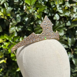 May include: A gold-toned tiara with a pointed center and a curved band. The tiara is encrusted with numerous clear, sparkling rhinestones. The tiara is displayed on a mannequin head against a backdrop of green foliage.