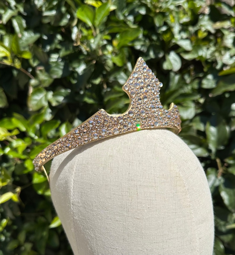 May include: A gold-toned tiara with a pointed center and a curved band. The tiara is encrusted with numerous clear, sparkling rhinestones. The tiara is displayed on a mannequin head against a backdrop of green foliage.