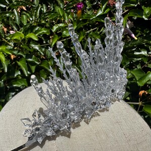 May include: A clear, crystalline tiara featuring tall, jagged icicle-like spikes and intricate snowflake-shaped accents at the base. The structure is adorned with silver glitter detailing, creating a reflective, icy appearance across the entire headpiece.