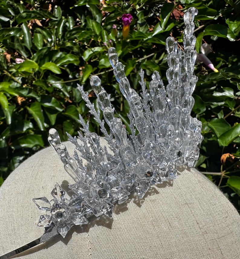 May include: A clear, crystalline tiara featuring tall, jagged icicle-like spikes and intricate snowflake-shaped accents at the base. The structure is adorned with silver glitter detailing, creating a reflective, icy appearance across the entire headpiece.