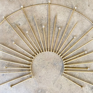 May include: A gold metal sunburst wall decor with a circular base and multiple pointed rays. Each ray is topped with a clear rhinestone.