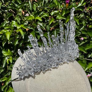 May include: A clear, crystalline crown featuring tall, jagged, icicle-like spikes and intricate, star-shaped snowflake accents. The structure is embellished with silver glitter, creating a textured, reflective surface that mimics frozen ice.