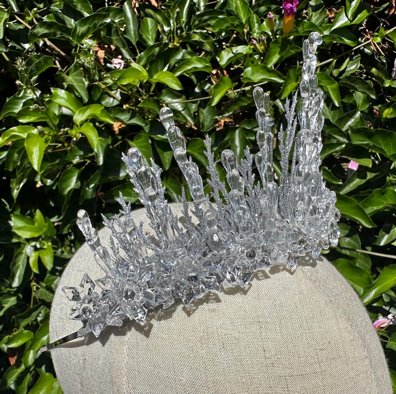 May include: A clear, crystalline crown featuring tall, jagged, icicle-like spikes and intricate, star-shaped snowflake accents. The structure is embellished with silver glitter, creating a textured, reflective surface that mimics frozen ice.