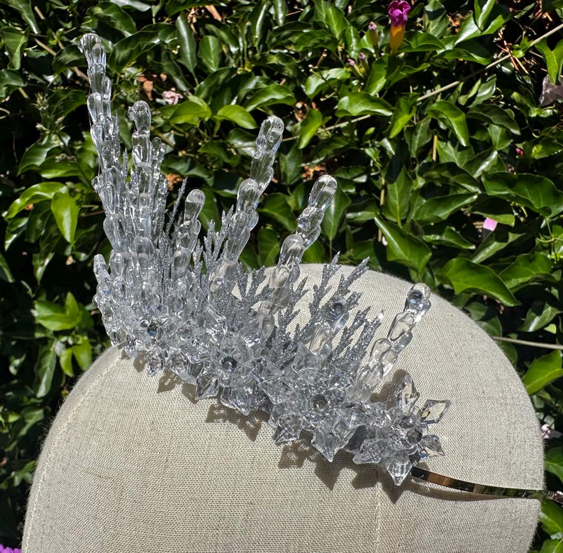 May include: A clear, crystalline crown featuring tall, icicle-like spikes and intricate, star-shaped snowflake accents. The structure is covered in a fine, silver glitter finish, creating a textured, sparkling appearance across the entire headpiece.