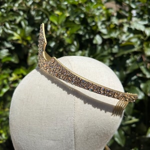 May include: A gold-toned tiara with a curved design, encrusted with small, sparkling crystals. The tiara is set on a mannequin head, showcasing its shape and detail. The background features lush green foliage.