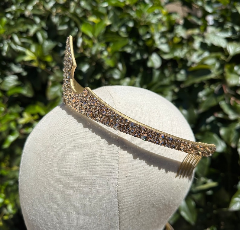 May include: A gold-toned tiara with a curved design, encrusted with small, sparkling crystals. The tiara is set on a mannequin head, showcasing its shape and detail. The background features lush green foliage.