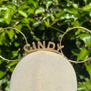 Puede incluir: Una diadema dorada con el nombre "Cindy" escrito en letras brillantes. La diadema tiene dos grandes acentos circulares que se asemejan a orejas. El fondo es un exuberante follaje verde.