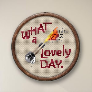 May include: A round, wooden-framed embroidery hoop with a white fabric background. The fabric has a red embroidered message that reads "What a lovely day." There is a small black and white embroidered image of a cat with a red and orange embroidered flame coming out of its mouth.