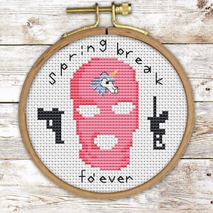 May include: A white embroidery hoop with a pink and black cross-stitch design. The design features a pink balaclava with a unicorn on top, two guns on either side, and the text "Spring break forever".