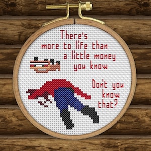 May include: Cross-stitch embroidery hoop with a white background and red text that reads "There's more to life than a little money you know Don't you know that?" The embroidery also features a pixelated image of a person in a red shirt and blue jeans.