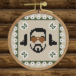 May include: A cross-stitch embroidery hoop with a green and white border. The center of the hoop features a brown-skinned person with a black beard and a brown mustache. The person is wearing a brown hat and a brown shirt. The background is a wooden wall.