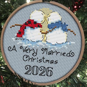 Może przedstawiać: Okrągły drewniany tamborek z haftem krzyżykowym przedstawiającym dwa bałwany w czerwonym i niebieskim szaliku. Napis A Very Married Christmas 2026 jest wyhaftowany czarną nicią na jasnoniebieskim tle tkaniny wewnątrz ramy.