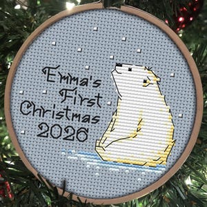 May include: A circular wooden embroidery hoop containing a cross-stitch design of a white polar bear sitting on blue ice. The text reads Emma's First Christmas 2026 in black lettering against a light blue background with small white squares.