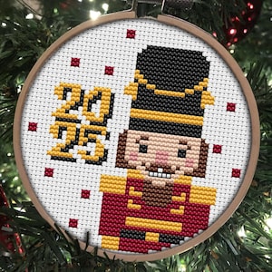 May include: A white cross-stitch ornament with a red and yellow nutcracker design and the text "2015" in yellow thread. The ornament is in a wooden hoop.