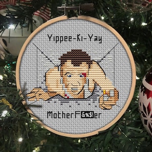 Peut inclure: Une décoration de Noël en point de croix gris et blanc représentant une image pixélisée d'un homme avec le texte "Yippee-Ki-Yay MotherF%&*!er".
