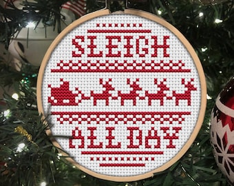 May include: A white circular ornament with red cross-stitch embroidery that reads "Sleigh All Day". The ornament is hanging on a green Christmas tree.