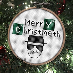 May include: A white circular ornament with a green and black cross-stitch design. The ornament features the text "Merry Christmas" and a stylized image of a man wearing a black hat and sunglasses. The text "C" and "Y" are in green squares with the numbers "6" and "39" in the top right corner of each square.