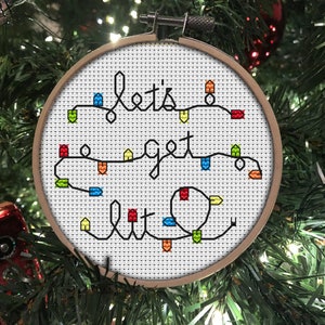 A 4inch, round, cross stitch Christmas tree ornament that spells Lets Get Lit with a string of colourful fairy lights.