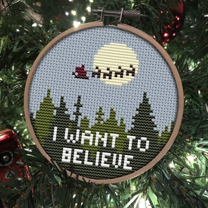 Könnte beinhalten: Ein Kreuzstich-Anhänger mit blauem Himmel, gelbem Mond und einem roten Schlitten, der durch die Luft fliegt. Der Anhänger hängt an einem grünen Weihnachtsbaum und trägt die Aufschrift "I WANT TO BELIEVE!" in weißen Buchstaben.