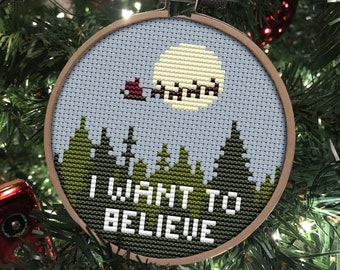 May include: A cross-stitch ornament with a blue sky, a yellow moon, and a red sleigh flying through the air. The ornament is hanging on a green Christmas tree and says "I WANT TO BELIEVE!" in white letters.