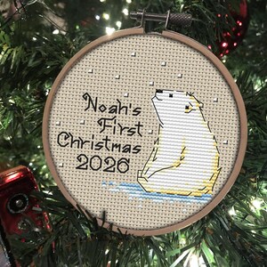 May include: A circular wooden embroidery hoop containing a cross-stitch design of a white polar bear sitting on blue ice. The text reads Noah's First Christmas 2026 in black lettering, with small white squares representing falling snow.
