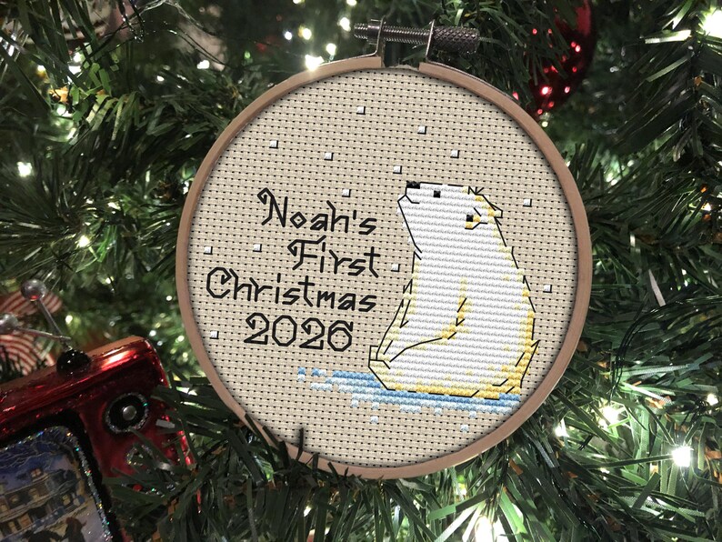 May include: A circular wooden embroidery hoop containing a cross-stitch design of a white polar bear sitting on blue ice. The text reads Noah's First Christmas 2026 in black lettering, with small white squares representing falling snow.