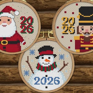 May include: Three circular wooden embroidery hoops containing cross-stitch patterns of a Santa Claus, a nutcracker, and a snowman. Each design features the year 2026 stitched in distinct colors against a light beige fabric background.