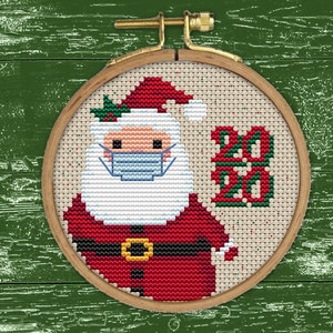 以下が含まれることがあります： サンタクロースがマスクを着けて「2020」の文字が書かれたクロスステッチの模様。模様は木製の輪で囲まれています。