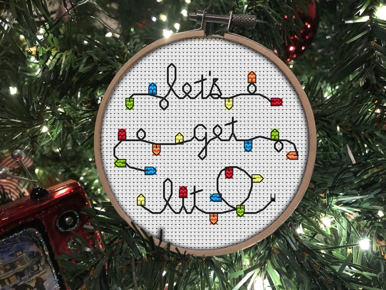 A 4inch, round, cross stitch Christmas tree ornament that spells Lets Get Lit with a string of colourful fairy lights.